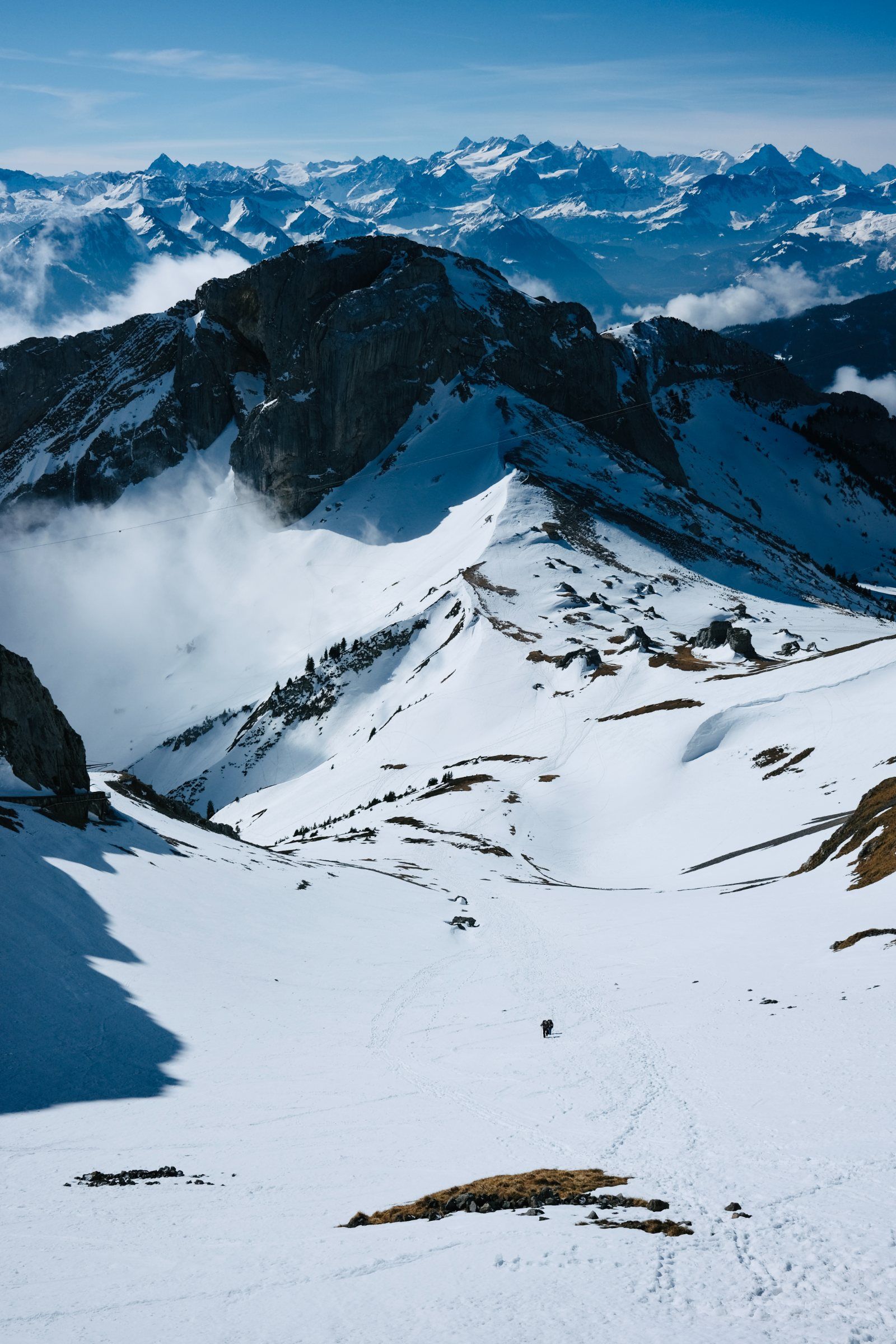 Snowfield below an alpine ridge, with a distant range of peaks above the clouds